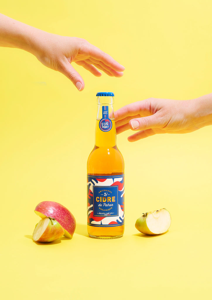 La boisson artisanale de Le Coq Toqué!, cidre brut du patron bio, petit format de 33cl, sur un fond jaune avec des pommes découpés et deux mains qui attrapent le cidre 